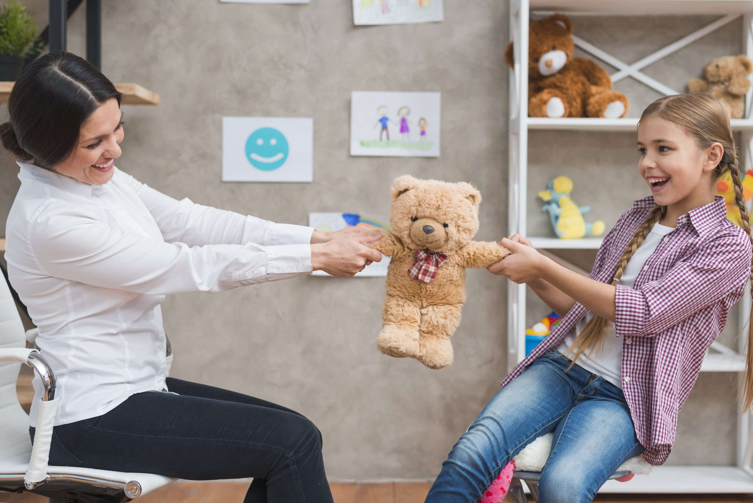 Female psychologist and smiling girl sitting face to face pulling the teddybear